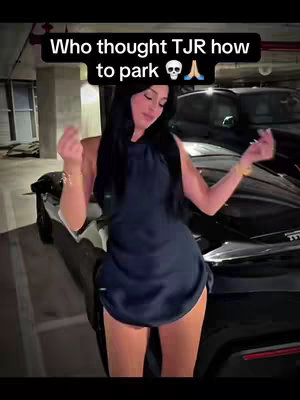 A person stands in front of a black car in a parking garage. The person is wearing a short, dark blue dress and posing. Text overlayed on the image reads Who thought TJR how to park.