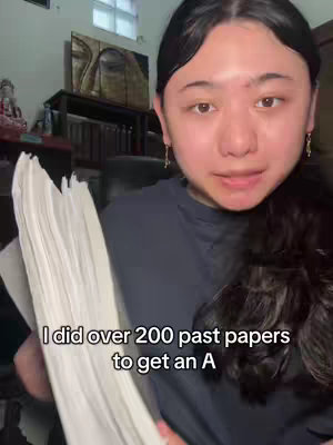 A young woman with long dark hair is holding a large stack of white papers. She's looking at the camera and speaking. The background is somewhat blurred, showing a room interior with a painting, a table, and other objects. Text overlayed on the image reads, 'I did over 200 past papers to get an A.'