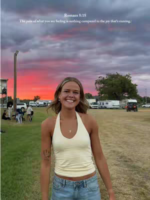 A young woman with blonde hair smiles at the camera, wearing a cream-colored halter top and denim shorts, standing in a sunny outdoor setting. The background features a vibrant sunset sky, with a field and hints of parked vehicles and people in the distance.