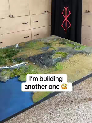 A detailed diorama of a fantasy landscape, possibly representing Middle-earth or a similar fictional setting. The diorama features mountainous terrain and a body of water, with varying shades of green, brown, and blue for the landscape and water, respectively. A black curtain with a red emblem hangs in the background.