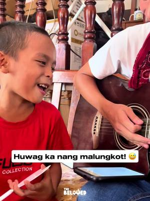 A young boy with short hair is smiling widely while seated next to a person, possibly an adult, who is playing the guitar. The boy is wearing a red short-sleeved shirt. The guitar player is wearing a white shirt covered by a red bandana. The cover image showcases a musical setting.