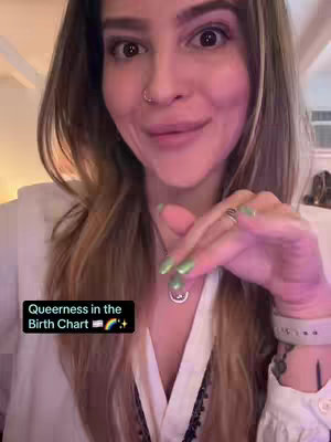 A woman is seen from the chest up, facing the camera directly. She has long, light brown hair with highlights. She's wearing a white top. Her face is slightly blurred, with a soft focus, but it's clear that she is smiling slightly. Overlaying her body is a black box with the words "Queerness in the birth chart" and the rainbow emoji and transgender symbol. The background is softly lit, suggesting an interior setting.