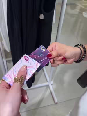The image shows two hands, both with manicured nails, holding up two credit cards. One card has a swirl design with pastel colors, while the other is dark with a constellation design. The background is blurred, but appears to be a clothing store or similar retail setting.