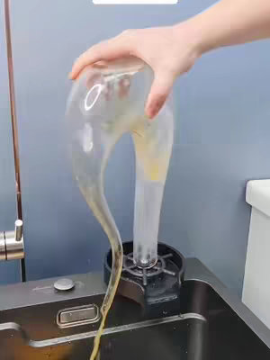 A hand holds a transparent, undulating object filled with a pale yellow liquid, pouring it into a black circular device set in a dark sink.