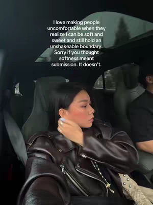 A woman is shown from the shoulders up, in the front seat of a car. She is looking towards her right and slightly down, with her right hand touching her left clavicle. She's wearing a brown leather jacket and her skin has visible makeup. Beside her, a person in a black shirt is visible. The background is blurred but visible - a car interior with a skylight and outdoor trees.