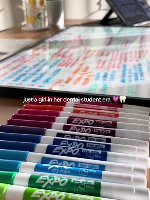 The image shows a collection of colored Expo dry-erase markers neatly arranged in a row. Above the markers, a blurred background showcases a whiteboard with colorful notes and diagrams, suggesting a study or learning environment. The text overlayed on the image reads "just a girl in her dental student era," along with a heart and a tooth emoji.