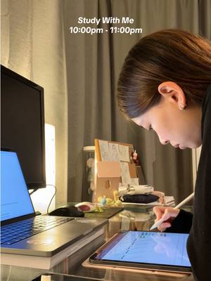A woman is shown in a dimly lit room, studying. She is sitting at a desk with a laptop, an open tablet, and a pen in hand. The background includes a curtain, a wall, and some items on the desk, with soft light creating a warm atmosphere.