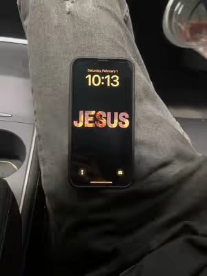 The image shows a person's legs and a smartphone resting on them. The phone's screen displays the time 10:13, the word 'JESUS' in large, stylized font over a blurry, fiery background, and the date is 'Saturday, February 1'. The leg is wearing gray jeans with a tear. A partially visible cup is in the upper right corner.