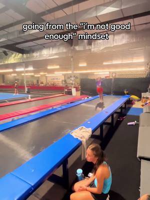 A large indoor gymnastics gym with various equipment. In the foreground, a woman in a blue tank top is sitting with a water bottle. In the background, a gymnast wearing red performs a tumbling routine. The text overlay is white, placed at the top, while the rest appears naturally in the background.