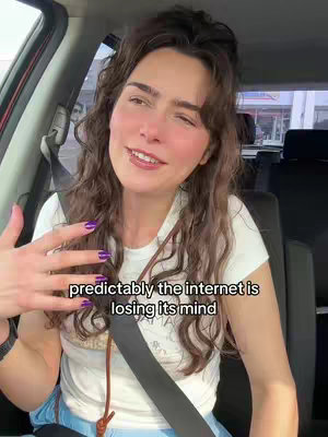 A woman with long, wavy brown hair, is seated in a car with an visible seatbelt. She is wearing a white t-shirt and has long purple nails. The text overlay reads, 'predictably the internet is losing its mind'. Her expression and pose suggest she is speaking or reacting to something.