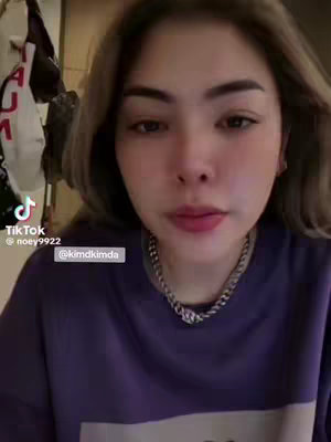 A selfie-style video cover featuring a woman with light brown hair and a purple shirt. She is positioned in the center, looking directly at the camera with a neutral expression. A silver chain necklace is visible. The background is a bit blurred or out of focus, suggesting an interior setting. Text overlays include the TikTok logo, username and a user tag at the bottom left, middle and middle right respectively.