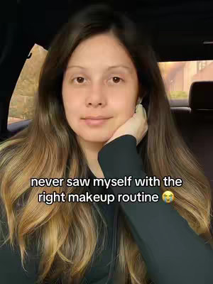 A woman with long, wavy brown hair poses inside a car, resting her hand under her chin. Her face is turned slightly towards the camera, showcasing a natural, makeup-free look. The background is blurred, suggesting an outdoor setting. Text overlayed on the image reads, 'never saw myself with the right makeup routine' and includes a crying face emoji.