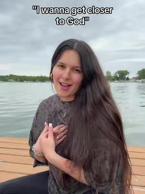 A woman with long dark hair smiles while sitting on a dock. The background shows a lake with trees and a cloudy sky.