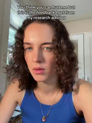 A young woman with curly brown hair and blue eyes is looking directly at the camera with a slightly annoyed expression. She's wearing a blue tank top and a necklace with a pendant. The background is slightly blurred, suggesting an indoor setting with natural light.