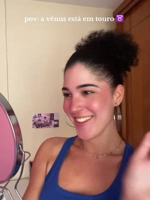 A person in a blue tank top smiles while looking in the direction of a pink mirror. The person has dark, curly hair and a light complexion, with blush visible on their cheeks. Behind them, the background is a light-yellow wall. A sign with pictures is slightly visible to the left.