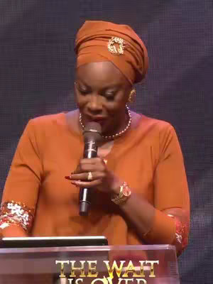 A medium shot shows a Black woman giving a sermon with a microphone in hand. She wears an orange dress and a brown headwrap with an ornate brooch. Behind her is a screen that says 'THE WAIT IS OVER' and has a purple background.