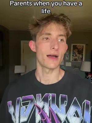 A young man with light brown hair is the central focus, framed from the chest up. He stands inside a room, appearing in front of a wall and various objects. His hair is styled with some volume and the look on his face suggests he's in mid-sentence. The background includes a soft, neutral-toned wall, with art. He is wearing a dark graphic t-shirt. The top third of the frame has the text 'Parents when you have a life'. The lighting is natural, suggesting daylight, and the overall tone of the image is casual and direct.