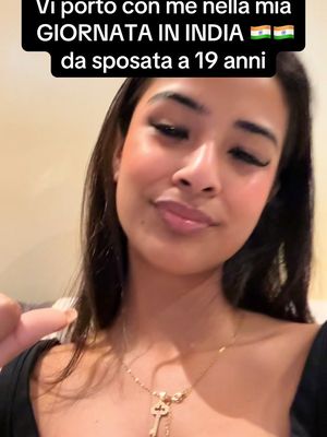 A selfie-style shot features a young woman with long, dark hair and a golden necklace. She is positioned slightly off-center with a soft, warm setting in the background. Her expression is gentle, and she appears lit from above, which gently highlights her features. Above her head, a text overlay in a bold font reads, "Vi porto con me nella mia GIORNATA IN INDIA ๐ฎ๐ณ๐ฎ๐ณ da sposata a 19 anni".