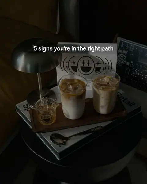 A low-angle shot of a wooden side table with two iced lattes, a glass carafe with honey, and a silver lamp. A white graphic with a smiling face logo sits behind the drinks. The color palette involves beige, brown and dark tones.