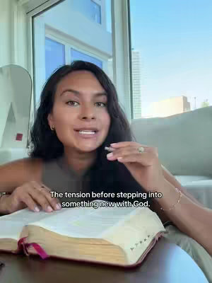 A woman is seated, holding an open book, with a window and city skyline visible behind her. The text overlay reads, 'The tension before stepping into something new with God.'