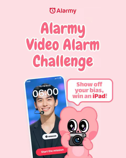 A promotional image for an alarm app, featuring the app interface displayed on a phone showcasing a smiling young man as the alarm image. The app is set to 6:00 AM. A pink cartoon character with a camera is smiling in the lower right corner, and speech bubble that says, 'Show off your bias, win an iPad!' The background is a soft pink gradient.