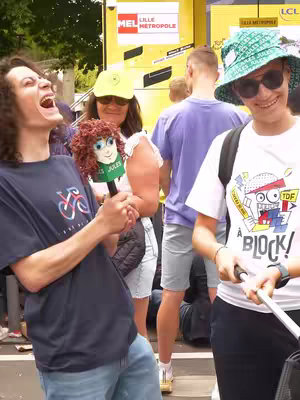 A group of people are gathered outdoors. The main subject on the left is a young man in a dark blue t-shirt, jeans, and with long brown curly hair, he is laughing with his mouth wide open. He is holding a microphone with a caricature head of what appears to be a character. To the right, there is a woman wearing a hat and sunglasses, and a man with a purple t-shirt in the background. The background features a large yellow structure.