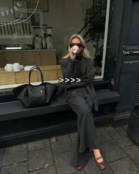 The cover image shows a woman sitting on a black bench outside a cafe. She is wearing a dark blazer and pants, paired with dark sunglasses, holding a coffee cup up to her mouth. A large black tote bag sits to her left. The background has a window display showing coffee-making equipment with the word "COFFEE" on the window.