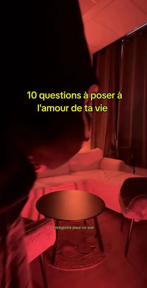 The image shows a person angled downwards, partially obscuring the view with a dark, undefined form in front of a room with soft, indirect red lighting. The scene includes a white sofa, a round black table, and muted chairs. Text overlays appear in a yellow font, posing questions.