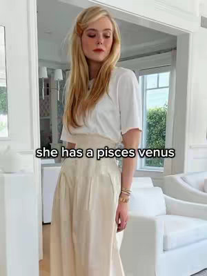 A woman with blonde hair stands in a well-lit room, wearing a white t-shirt and a cream-colored skirt. Text overlayed on the image reads, "she has a Pisces venus".