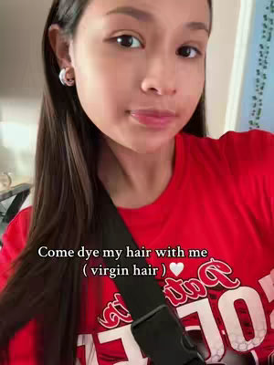 A young woman with long dark hair and hoop earrings smiles at the camera while holding some of her hair. She is wearing a red top with white text on it and a black cross-body strap. Text overlayed on the video says, "Come dye my hair with me (virgin hair)."