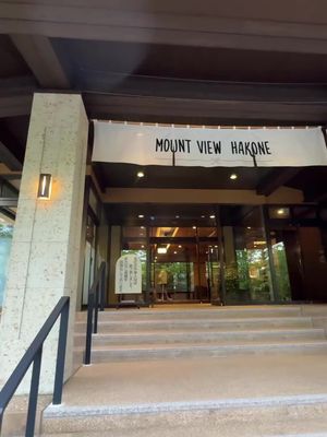 The image showcases the entrance of a building, likely a hotel or resort, under an overcast sky. The facade is predominantly light-colored, possibly beige or tan, with several steps leading up to glass doors. A banner that reads "MOUNT VIEW HAKONE" is hung above the entrance. The entrance is flanked by architectural columns and features a combination of stone and darker wooden accents. The steps are lit by an array of lights, and a sign stands to the side of the entrance offering some basic information. The overall composition is symmetrical, conveying a sense of order and grandeur.