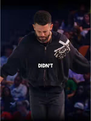 A man stands in a large space, appearing to be a stage or event area, with a crowd in the blurry background. He is wearing a black zip-up hoodie with a graphic design, possibly a skull or other artistic element, on the side. The word "DIDN'T" is overlaid in white text on the lower part of the image.
