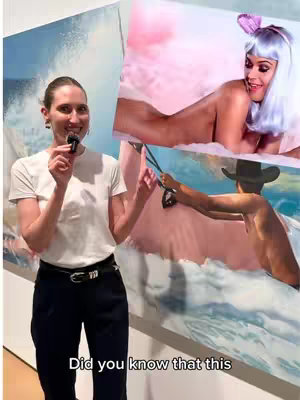 A woman stands in front of art, holding a microphone and pointing at a painting of Katy Perry.
