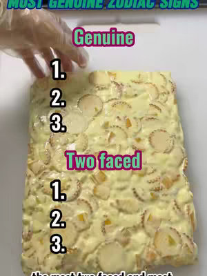 A tray of dessert is shown. The dessert consists of a yellow base, possibly custard or a similar substance, topped with circular, light-colored decorations. Text overlays rank the signs. A hand wearing a glove appears to the left, touching the dessert.