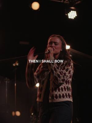 A medium shot features a woman on stage, illuminated by stage lights. She is singing into a microphone, her face conveying emotion and focus. The background is dimly lit, with stage equipment visible. Text overlayed on the image reads "THEN I SHALL BOW". The woman wears a patterned cardigan.
