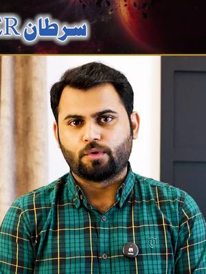 A man with dark hair and a beard is centered in the frame, wearing a green and black plaid shirt. The background has a dark neutral tone, with a blurred visual element at the top that includes shades of red, orange, and blue, like a planetary body. Text in Arabic is written above the man's head. A small circular pin with a logo is attached to his shirt.