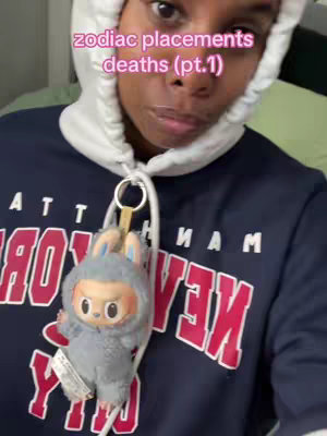 The cover image shows a person, likely the creator, wearing a navy blue hoodie with a New York graphic. The person is wearing a white hood over their head, and has an accessory hanging from the zipper, a plush toy.  Text overlayed on the image reads "zodiac placements deaths (pt.1)" written in pink.  The person is looking toward the camera.