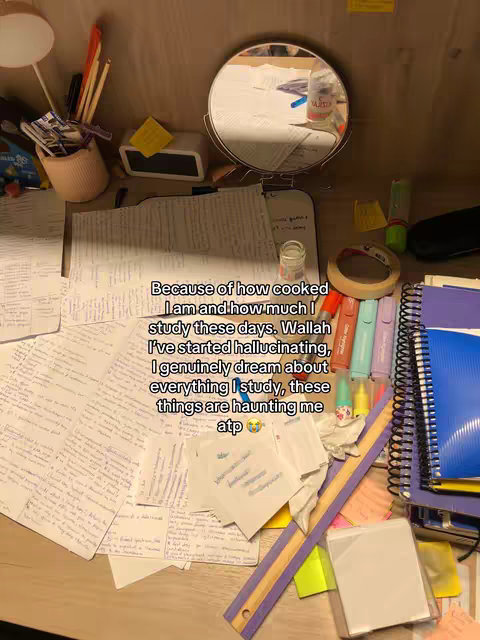 A top-down shot of a messy desk covered in papers, highlighters, notebooks, and small items. A mirror in a round frame is near the top of the frame. Additional items include pens, a lamp, and a timer. The color palette is a mix of warm and cool tones with a lot of white and yellow, and blue.
