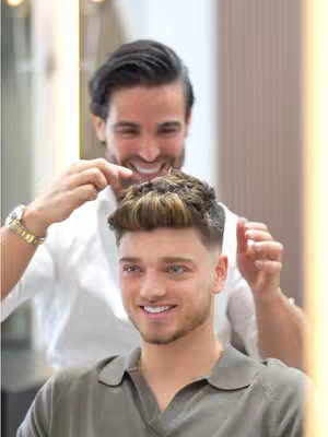 The image features two men. One man is in the foreground, looking directly at the camera with a smile. He is wearing a gray collared shirt. The other man is positioned behind him, looking at the man's hair and is smiling. The setting is a salon environment, and the overall composition focuses on a grooming or hairstyling theme.