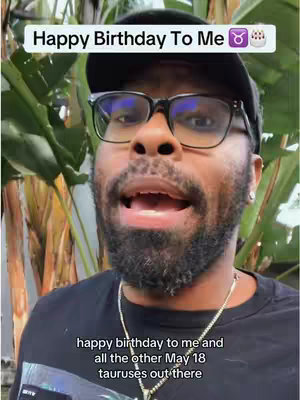 A man with a beard and glasses is the central focus of the image. He's wearing a black cap and t-shirt. White text on top says "Happy Birthday To Me" with a Taurus symbol and a cake emoji. The man is speaking, likely addressing the camera, and there's green foliage in the background with text at the bottom.
