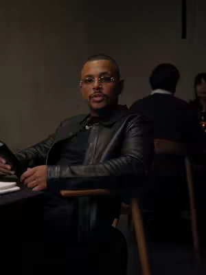 A man with glasses wearing a black leather jacket is seated at a table. Two people are seated in the background, slightly out of focus. The man is looking towards the camera. The lighting is dim.