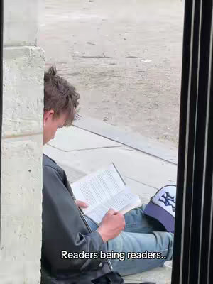 A person is sitting on the ground reading a book, with the frame partially obscured by a building's architectural element. The person is wearing a black jacket, blue jeans, and a baseball cap. The scene takes place outdoors.