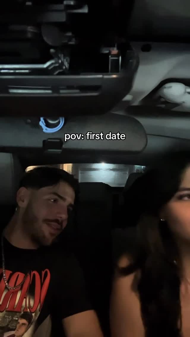 An interior car shot with a man and woman in the front seats, indicating a POV first date scenario. The man is turned towards the woman and smiling, suggesting a moment of connection.