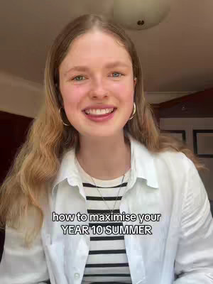 A young woman with long blonde hair smiles at the camera. She's wearing a white button-down shirt over a striped black and white top. The background shows a warm-toned room with a ceiling light.