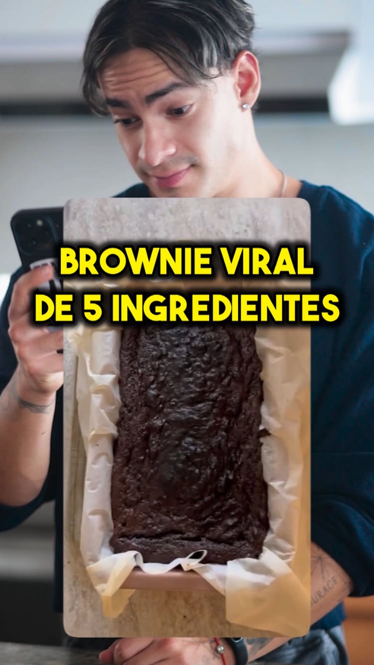 A man is holding a smartphone with the camera pointed at a baking dish of a brownie, a text overlay indicates the brownie is made with 5 ingredients. The man is looking down at the phone smiling.