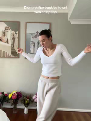 A woman wearing a white top and light grey sweatpants dances in a room. She is surrounded by flowers and artwork.