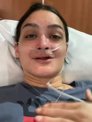 A close-up shot of a woman lying in bed, with a nasal cannula for oxygen. She is looking up and smiling. The background is a neutral, possibly clinical setting with white bedding.