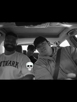 A black and white photo taken from inside a car. Three men are in the car, in the front seats, with one turned toward the camera with a smirk. The skull emoji is overlaid on the left.