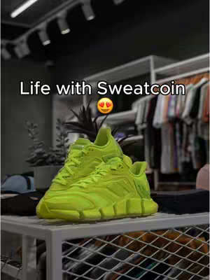 The image shows a pair of bright green athletic shoes on a white shelf. The shoes are the focal point. The background is blurred out, and includes a rack of clothes.