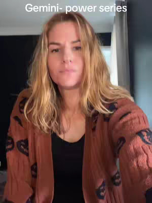A woman with blonde hair looks directly at the camera with a neutral expression. The background appears to be an interior scene. She's wearing a brown cardigan.
